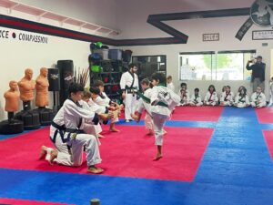 Group of children in white karate gis watching and practicing a kick in a dojo with red and blue mats and a line of training dummies along the wall likely during a class.