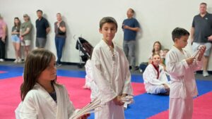 Children in white karate gis kneel on blue and pink mats, holding folded belts during class as spectators watch from the wall.