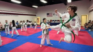 Group of children in white taekwondo uniforms with green belts practicing kicks on red and blue mats in a dojo.