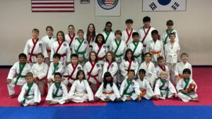 Group of children in white Taekwondo uniforms with colored belts posing for a class photo on matting, flags on the wall behind them.