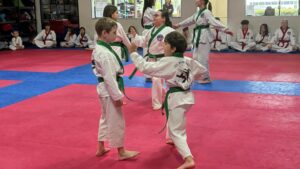 Two young taekwondo students in white uniforms with green belts high-five on a red-and-blue mat, with teammates seated along the wall behind them.