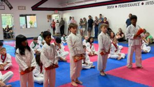 Group of children in white karate gi uniforms with colored belts on pink-and-blue mats during a class, some standing and some seated, with observers along the wall.