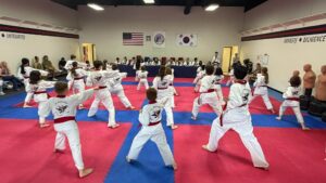 Group of children in white uniforms with red belts practicing martial arts in a dojo, facing outward in a wide stance on red-and-blue mats with spectators along the walls.