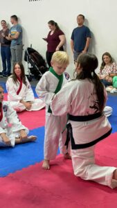 Children in karate uniforms kneeling on pink and blue mats while spectators stand along the wall in the background.