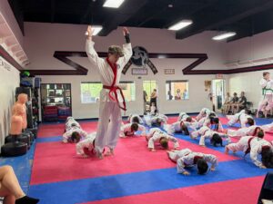 Instructor with red belt stands on pink/blue mat, arms raised as students in white gis crawl or bow during a karate class in a dojo.