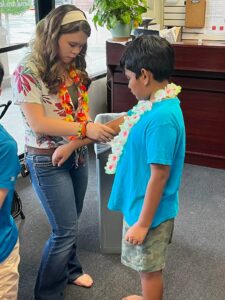 Teen girl in a floral shirt ties a bright lei around a boy's neck in a light-filled indoor space, barefoot on carpet.
