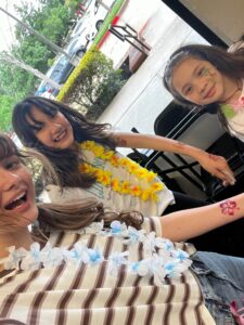 Three girls smile for a selfie, wearing yellow and blue flower leis and face paint in a sunny outdoor setting near a striped tablecloth.
