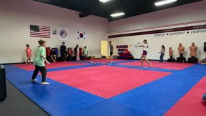 Group of kids and an instructor playing with small orange balls on a padded gym floor inside a martial arts dojo; flags hang on the wall.