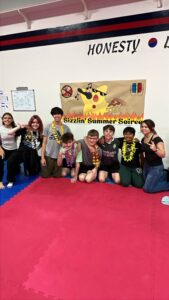 Group of kids posing for a photo in a gym, wearing colorful leis, in front of a 'Sizzlin' Summer Soiree' banner.