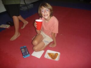 Child with braces and red glasses sitting on a red mat, smiling and holding a red cup; pizza slice on a plate nearby, a handheld game console and napkins on the floor.