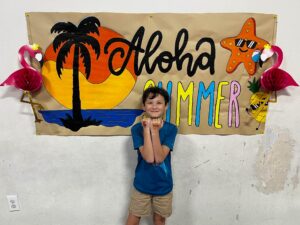 Smiling boy in a blue shirt stands in front of a tropical summer banner with a palm tree, starfish, and bright letters reading 'Aloha Summer.'