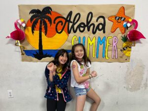 Three smiling girls posing in front of a tropical “Aloha Summer” banner with a palm tree, sun, starfish, and colorful decorations on a wall.