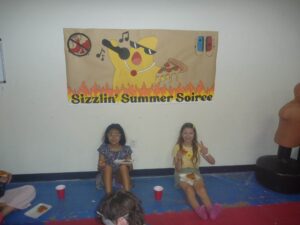 Poster of a yellow dog with sunglasses holding pizza, flames at the bottom, and the text "Sizzlin’ Summer Soiree" on the banner; people sit eating pizza in front of it.