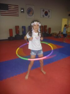 Young girl wearing a flower crown spins a colorful hula hoop in a gym.