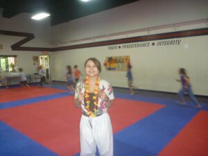 Smiling young woman in a tropical shirt and lei poses on red and blue gym mats; blurred children in background.