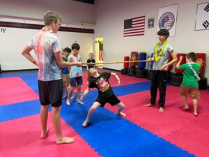 Children in a martial arts class hold a long wooden staff between them on a pink/blue padded floor, wearing leis and colorful shirts.