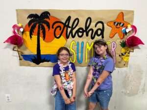 Two smiling girls wearing flower leis pose in front of a tropical luau backdrop with a palm tree and a starfish banner reading 'Aloha Summer'.