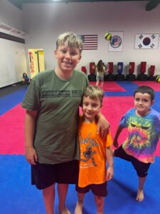 Three smiling kids pose together on blue and pink gym mats inside a martial arts dojo, with flag banners and practice punching bags in the background.