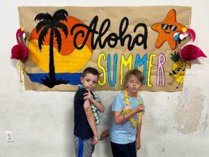 Two boys posing in front of a tropical summer banner with a palm tree, sun, starfish wearing sunglasses, and colorful lettering; they wear leis around their necks.