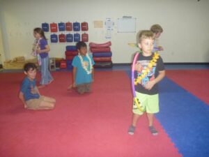 Children in a gym on red and blue floor mats, wearing leis; one boy holds a pool noodle while others stand nearby with a wall of red and blue padded bags in the background.