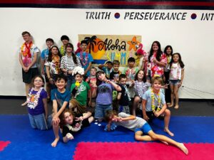Group of children wearing leis posing for a summer camp photo in a gym, with a tropical banner reading 'Aloha Summer' in the background.