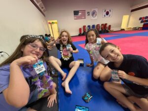 Kids in a martial arts gym pose for a selfie on blue and pink mats, wearing leis and holding name badges or cards.