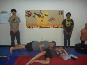 Kids at a summer party pose beside a 'Sizzlin' Summer Soiree' banner; several lie on foam mats on the floor nearby.