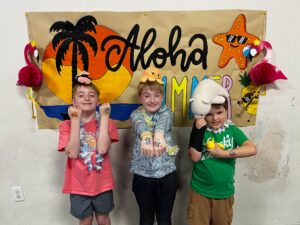 Three smiling kids in tropical party outfits pose in front of a beach banner with a starfish and palm tree decorations; hands show temporary tattoos.