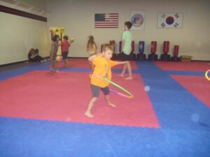 Children playing with colorful hoops on red-and-blue padded mats in a gym, with flags on the wall behind them (including the US flag).