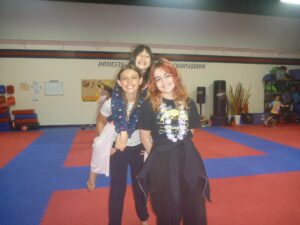Three smiling girls pose together in a gym with blue and red puzzle-piece mats and equipment in the background.