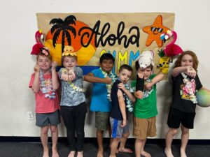 Group of children wearing leis and tropical accessories posing together in front of a beach-themed banner indoors.