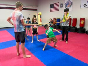 Children in a martial arts dojo practice with a long bamboo staff, watching as one student leans back in action on colorful mats.