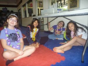 Four smiling children sit cross‑legged on red and blue foam mats indoors, wearing colorful flower leis.
