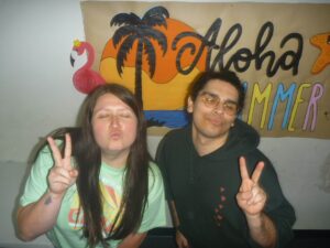 Two friends pose with peace signs and puckered lips in front of a colorful tropical mural with palm tree and bold lettering.