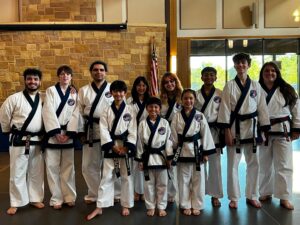 Group of smiling martial arts students in white uniforms with blue belts posing indoors, American flag in background.