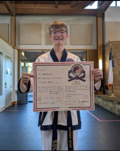 Young student in a martial arts uniform holds a certificate, smiling in a dojo with flags in the background.