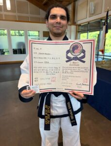 Man in a white martial arts gi with black belt holding a large certificate featuring a circular emblem and red-white-blue ribbon.