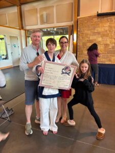 Young martial artist in a white gi holds a certificate, flanked by a smiling man and woman in a gym setting.