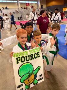 Four young taekwondo students with green belts smile while holding a large poster that says 'Green Turtle Force' and features a cartoon green turtle, in a gym with spectators in the background.
