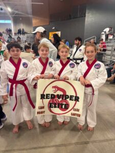 Four kids in white karate gis with red belts pose in a gym, holding a sign that says RED VIPER ELITE with names around the logo.