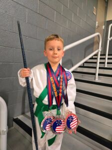 Young boy in a white karate gi with a green belt, wearing several medals, holding a blue staff in a stairwell.