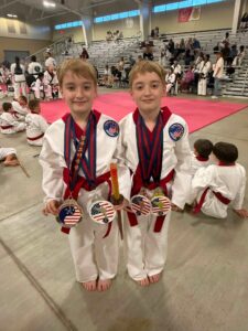 Two young boys in white and red taekwondo uniforms pose with multiple medals after a competition in a gym arena.