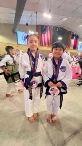 Two smiling children in white taekwondo uniforms with medals, posing after a competition in a gym with American flags in the background.