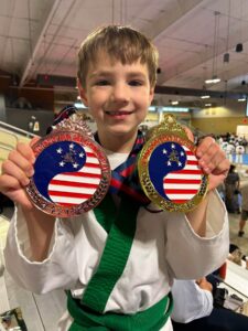 Smiling boy in a white martial arts gi with a green belt holds two large medals with American flag designs in a gym setting.