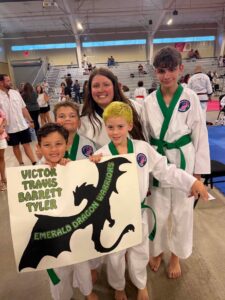 Four smiling children in white martial arts uniforms with green belts pose in a gym holding a poster with a black dragon silhouette and names on it.