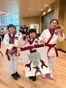 Three kids in karate uniforms wear medals and hold a poster at a trophy ceremony in a sunny indoor hall.