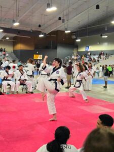 Female taekwondo competitor in a white gi with a blue belt delivering a kick on a pink competition mat, while spectators watch from the bleachers.