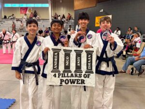 Four young martial artists in white uniforms with dark belts pose together in a gym, holding a banner that reads 'AND 4 PILLARS OF POWER' as spectators watch from the bleachers in the background.