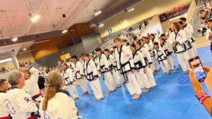 Group of children in white martial arts uniforms lined up on blue mats during a competition in a gymnasium with spectators in the stands.