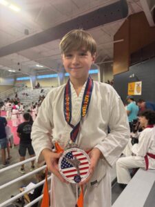 Smiling boy in a white karate gi with an orange belt, holding a patriotic trophy and medal at a competition.